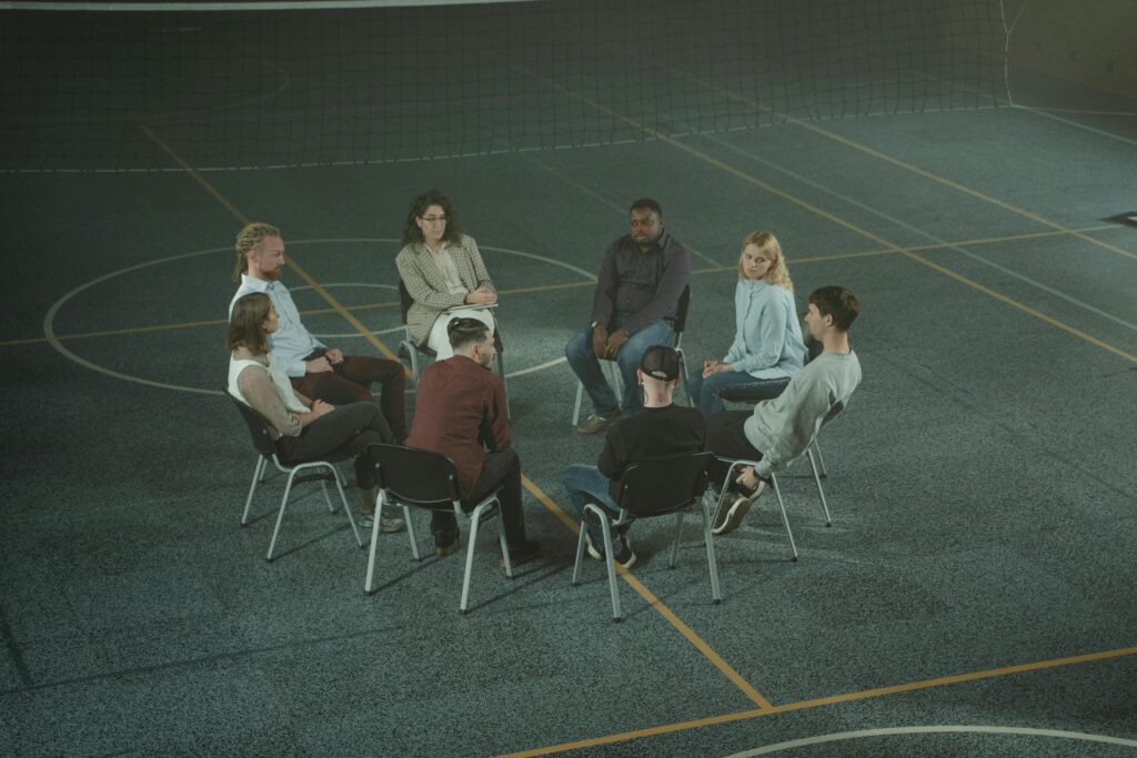A diverse group of people sitting in a circle during a therapy session in a sports hall.